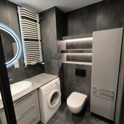 Marktor Marcin Poznański - Nowoczesna łazienka z szarą płytką imitującą kamień, podświetlane półki, umywalka nablatowa, pralka i grzejnik drabinkowy.