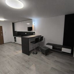 Marktor Marcin Poznański - Nowoczesne wnętrze w Sosnowcu: aneks kuchenny z barkiem i hokerami, szare panele podłogowe, białe ściany z czarnym panelem dekoracyjnym i szafka RTV.