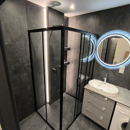 Marktor Marcin Poznański - Nowoczesna łazienka z czarną kabiną prysznicową, podświetlanym lustrem okrągłym, szarą umywalką nablatową z szafką i pralką Bosch.