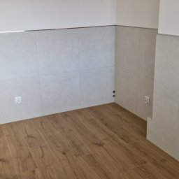 Marktor Marcin Poznański - Wnętrze po remoncie: jasne, kwadratowe płytki na ścianach, drewnopodobna podłoga, gniazdka elektryczne, listwa dekoracyjna na styku płytek i sufitu.