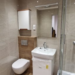 Marktor Marcin Poznański - Nowoczesna łazienka w jasnych beżach z drewnopodobną podłogą, wisząca toaleta, szafka z umywalką jeszcze w folii ochronnej i kabina prysznicowa z zaokrąglonym szkłem.