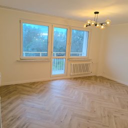 Marktor Marcin Poznański - Pusty pokój po remoncie z jasną podłogą ułożoną w jodełkę, białymi ścianami, oknami z widokiem na zieleń i nowoczesną lampą sufitową z kulistymi kloszami.