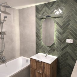 Marktor Marcin Poznański - Nowoczesna łazienka z zielonymi płytkami ułożonymi w jodełkę, podświetlanym owalnym lustrem nad drewnianą szafką umywalkową i białą wanną z chromowaną armaturą.