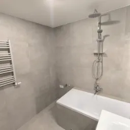 Wykończona łazienka w szarościach, z białą wanną i grzejnikiem drabinkowym; widoczny zestaw prysznicowy z deszczownicą i bateria wannowa.