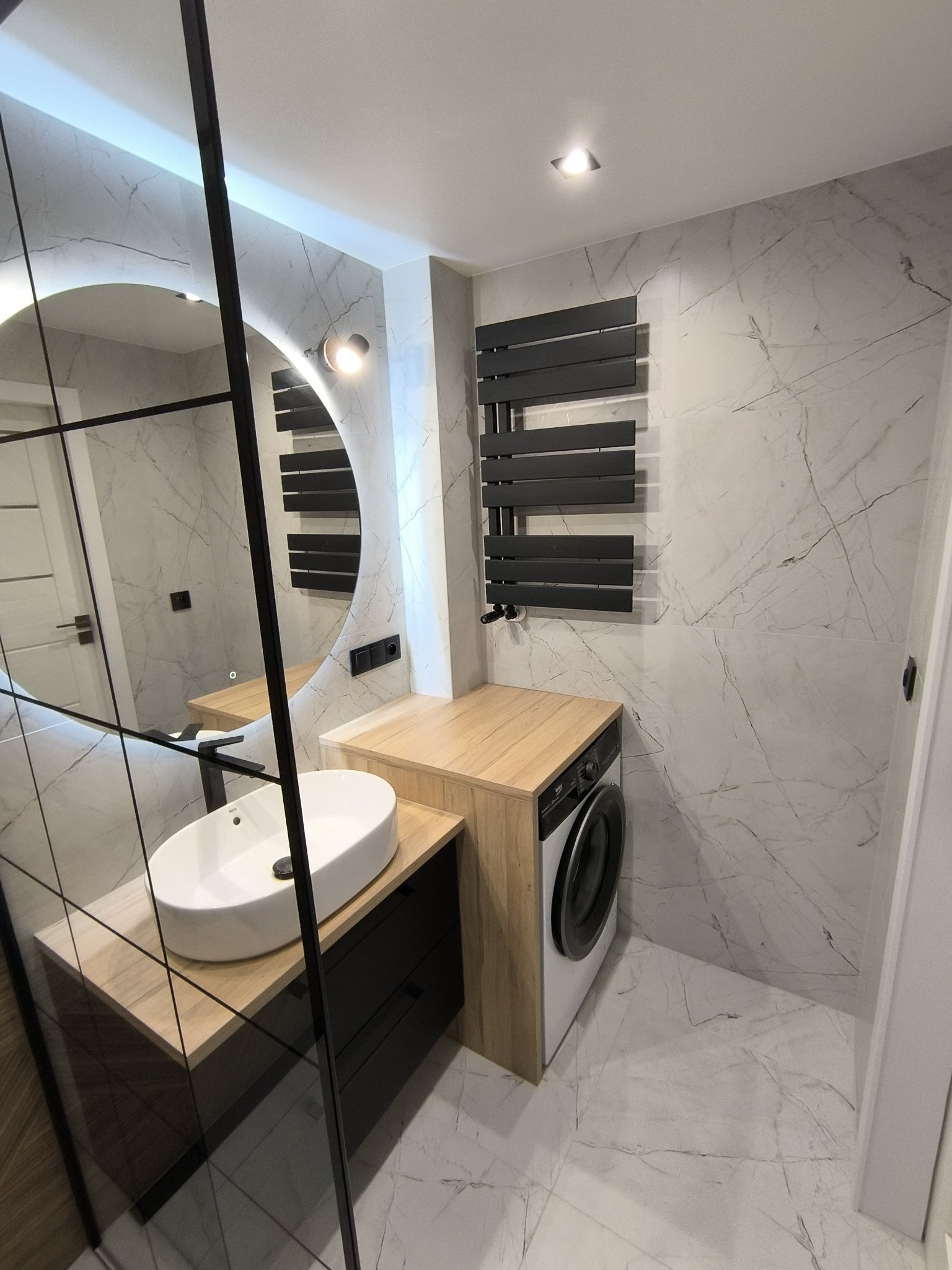 Nowoczesna łazienka z umywalką nablatową na szafce z szufladami, okrągłym lustrem z podświetleniem LED, grzejnikiem drabinkowym i pralką ukrytą pod blatem, wykończona płytkami imitującymi marmur.