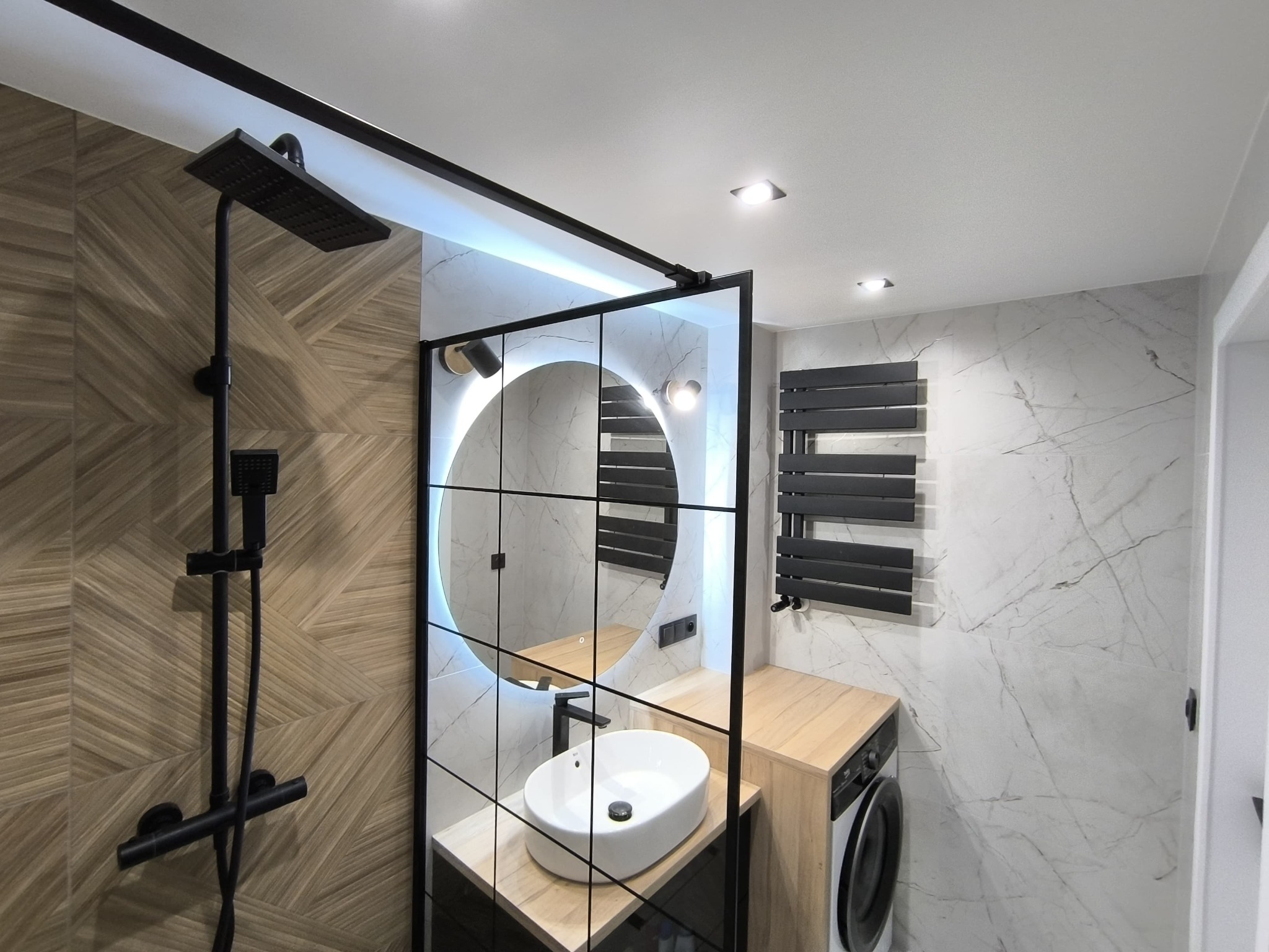 Nowoczesna łazienka z kabiną prysznicową typu walk-in, okrągłym lustrem z podświetleniem LED, umywalką nablatową, grzejnikiem drabinkowym w kolorze czarnym i pralką ukrytą pod blatem.