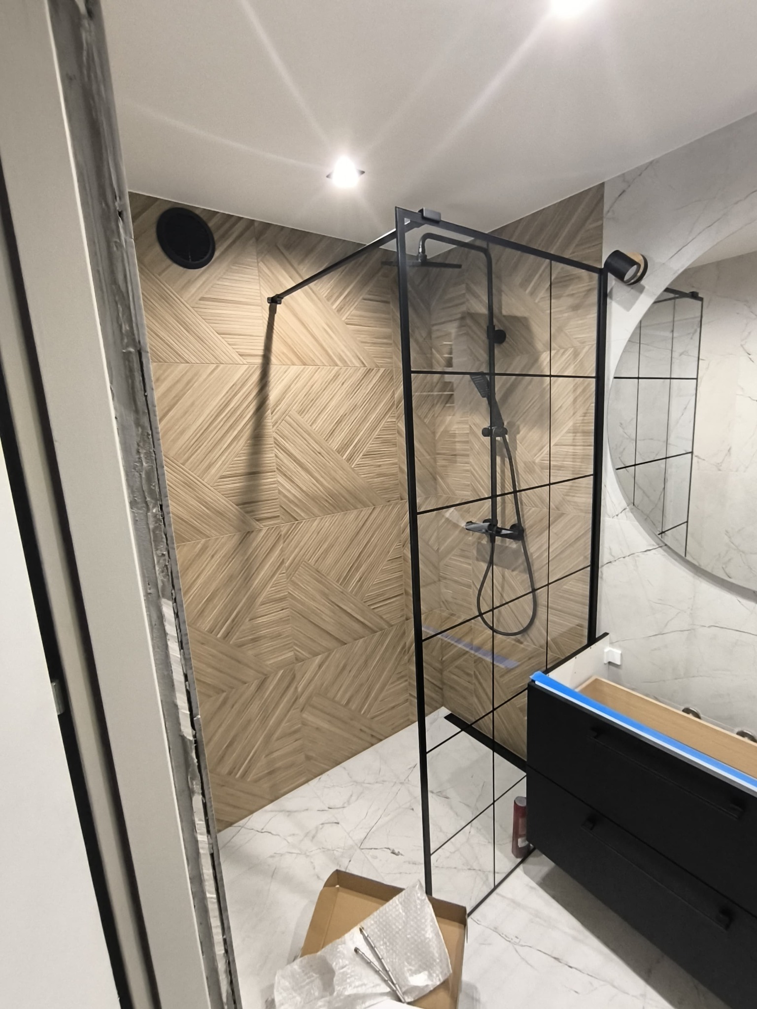 Montaż kabiny prysznicowej z czarnymi profilami i deszczownicą na tle ściany z drewnopodobnych płytek ułożonych w geometryczny wzór, widoczny fragment szafki pod umywalkę z niebieską taśmą malarską.