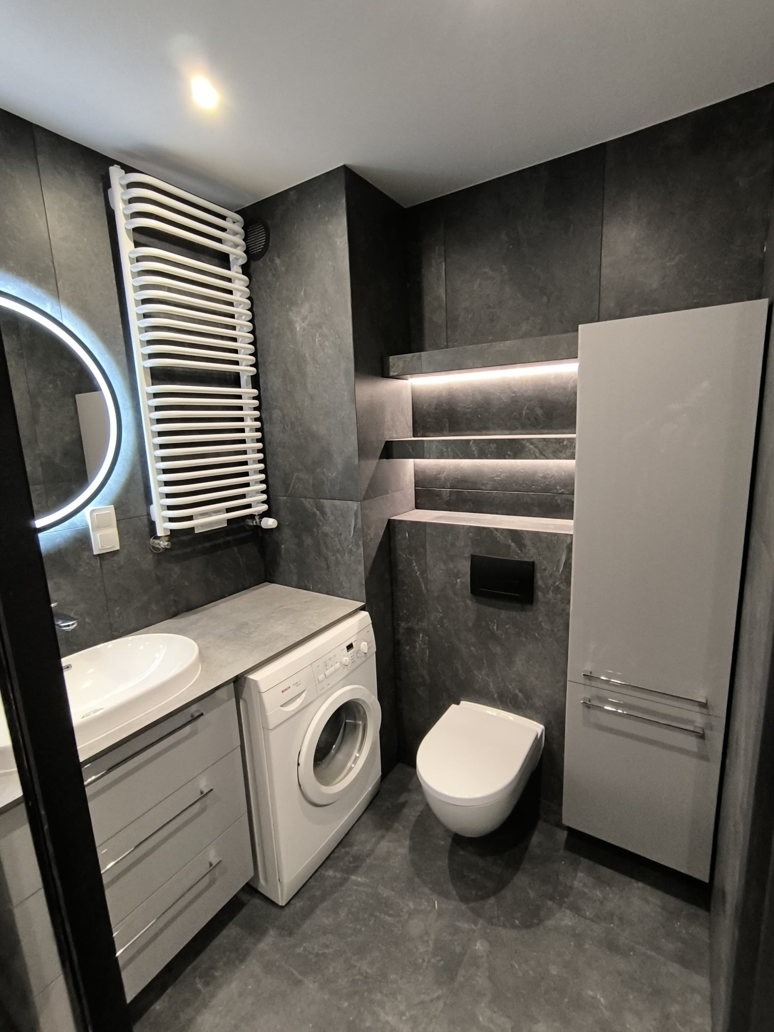 Nowoczesna łazienka z szarą płytką imitującą kamień, podświetlane półki, umywalka nablatowa, pralka i grzejnik drabinkowy.