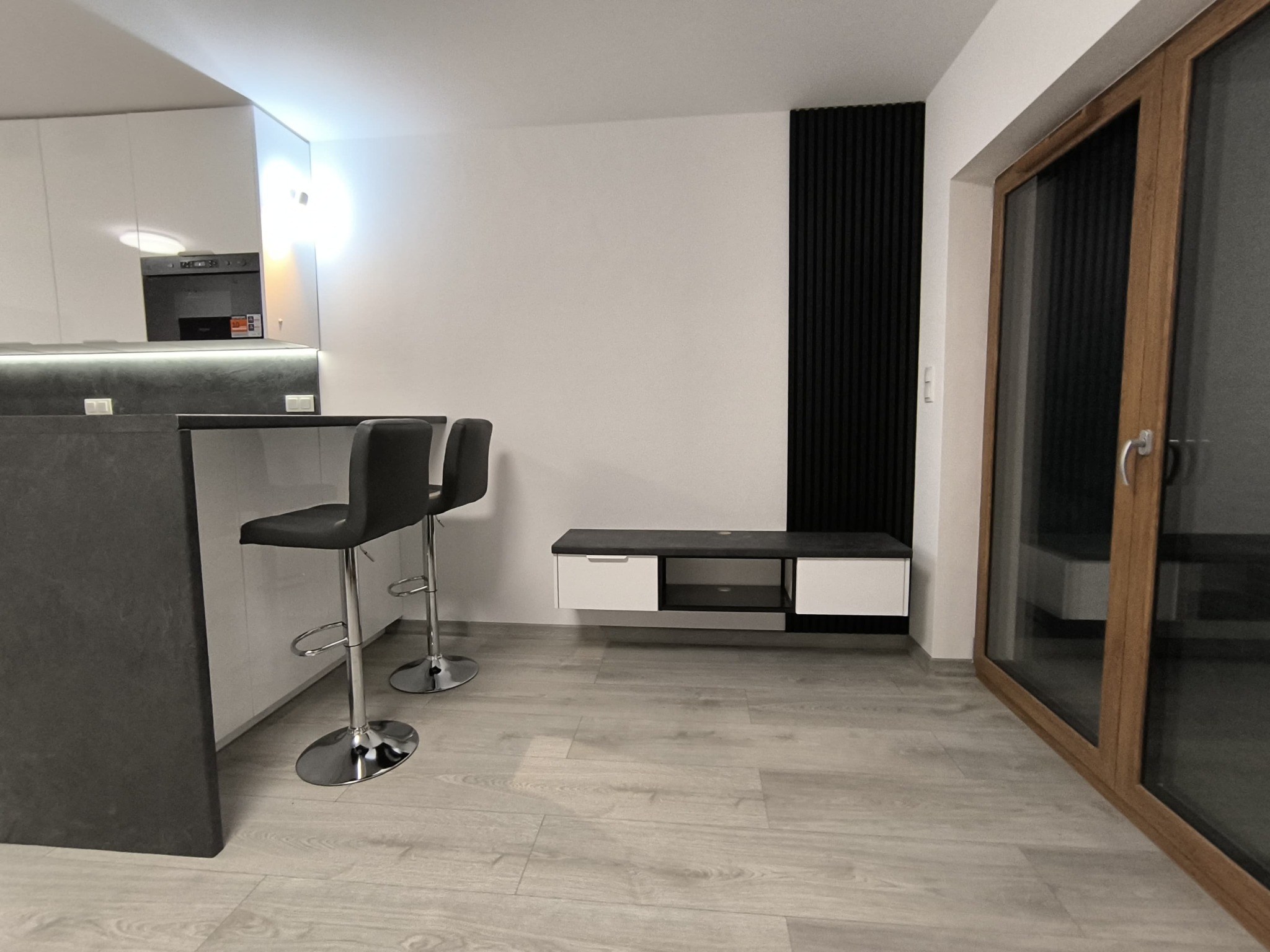 Nowoczesne wnętrze: minimalistyczna kuchnia z barkiem, hokerami, szafką RTV z czarnym panelem dekoracyjnym oraz drewnianą ramą okienną.