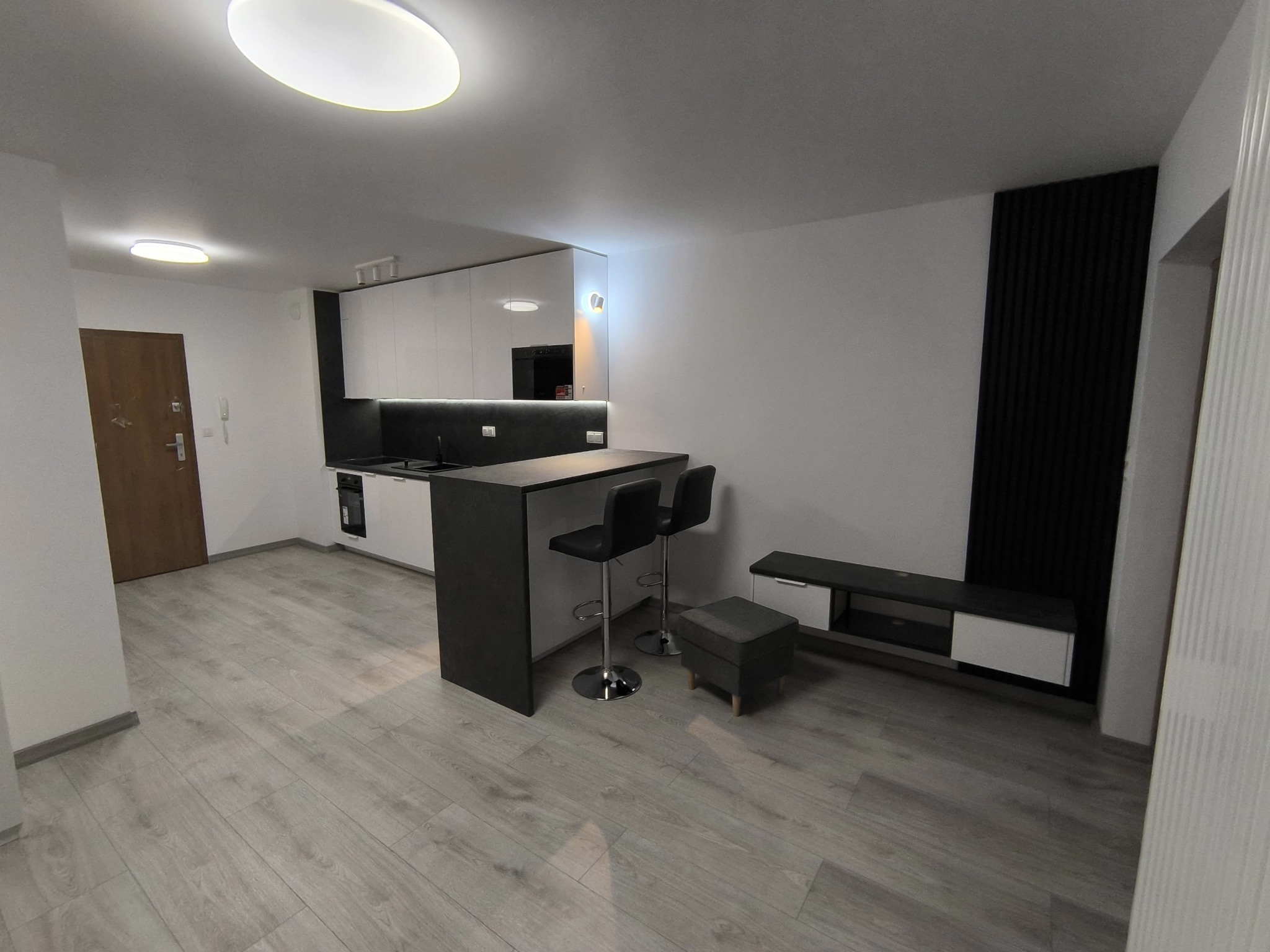 Nowoczesne wnętrze w Sosnowcu: aneks kuchenny z barkiem i hokerami, szare panele podłogowe, białe ściany z czarnym panelem dekoracyjnym i szafka RTV.