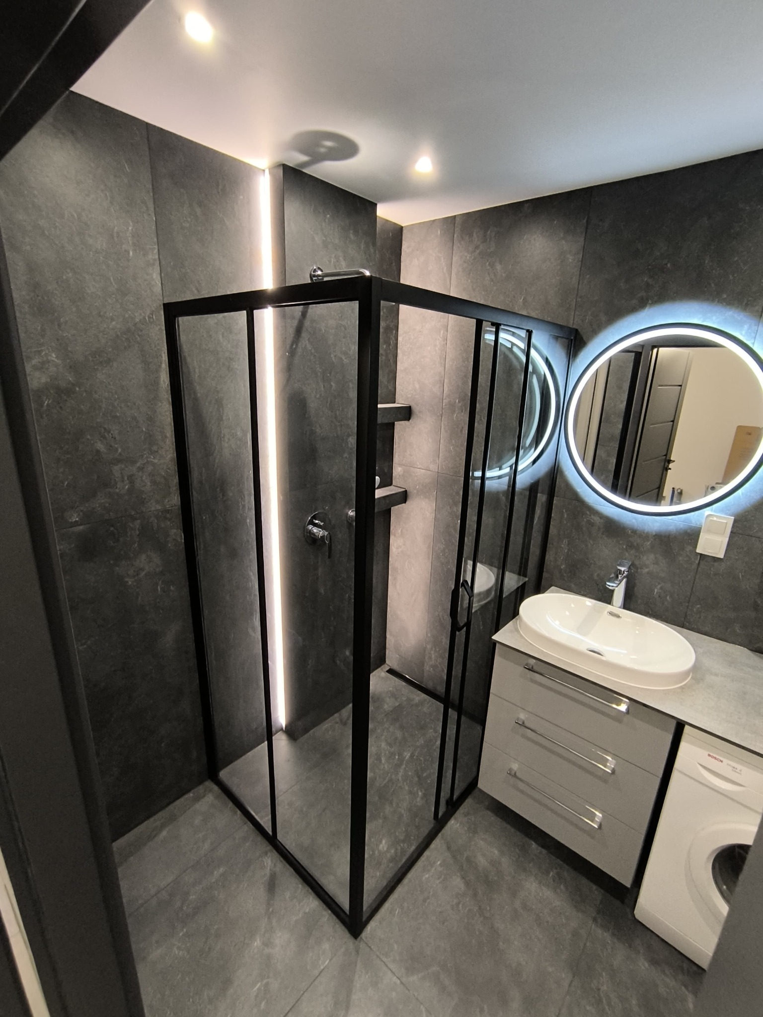Nowoczesna łazienka z czarną kabiną prysznicową, podświetlanym lustrem okrągłym, szarą umywalką nablatową z szafką i pralką Bosch.