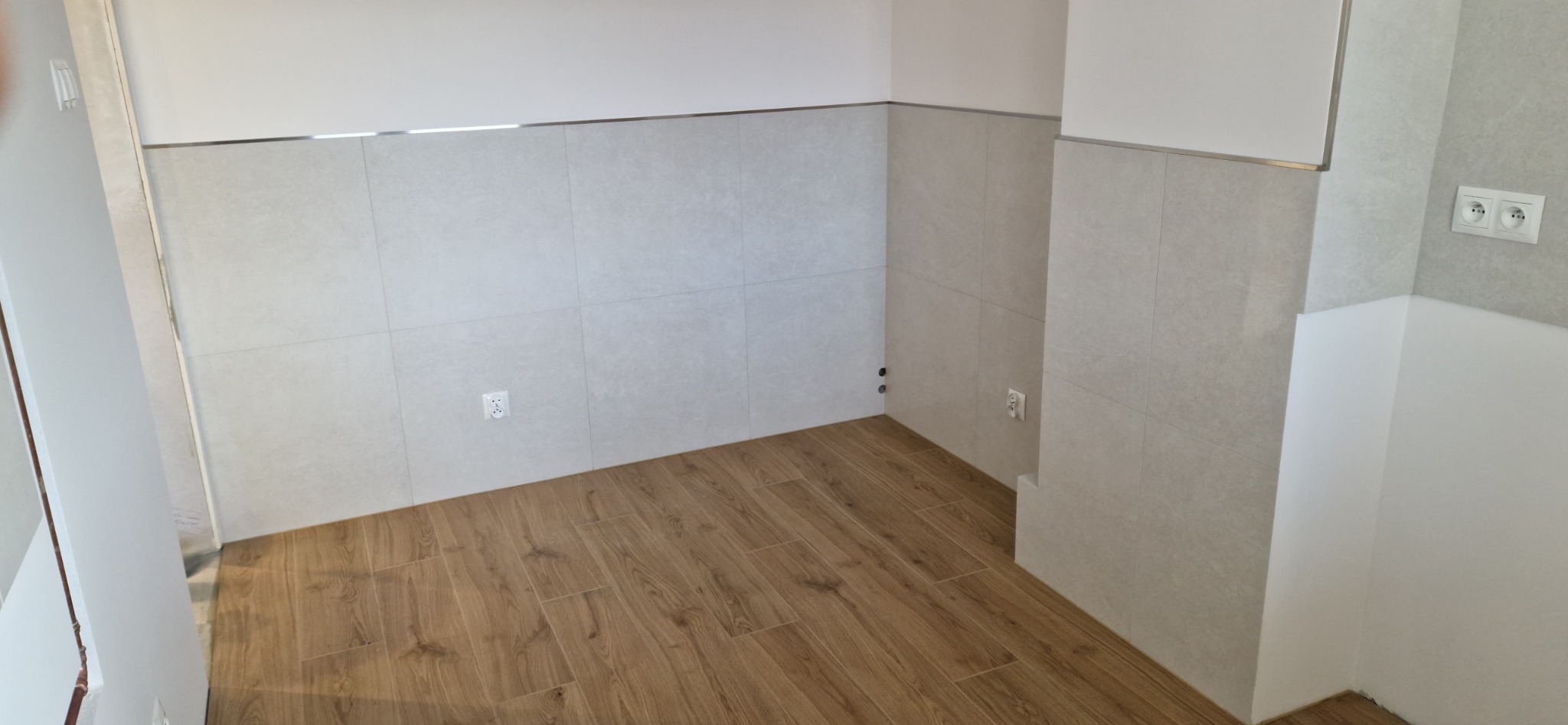 Wnętrze po remoncie: jasne, kwadratowe płytki na ścianach, drewnopodobna podłoga, gniazdka elektryczne, listwa dekoracyjna na styku płytek i sufitu.