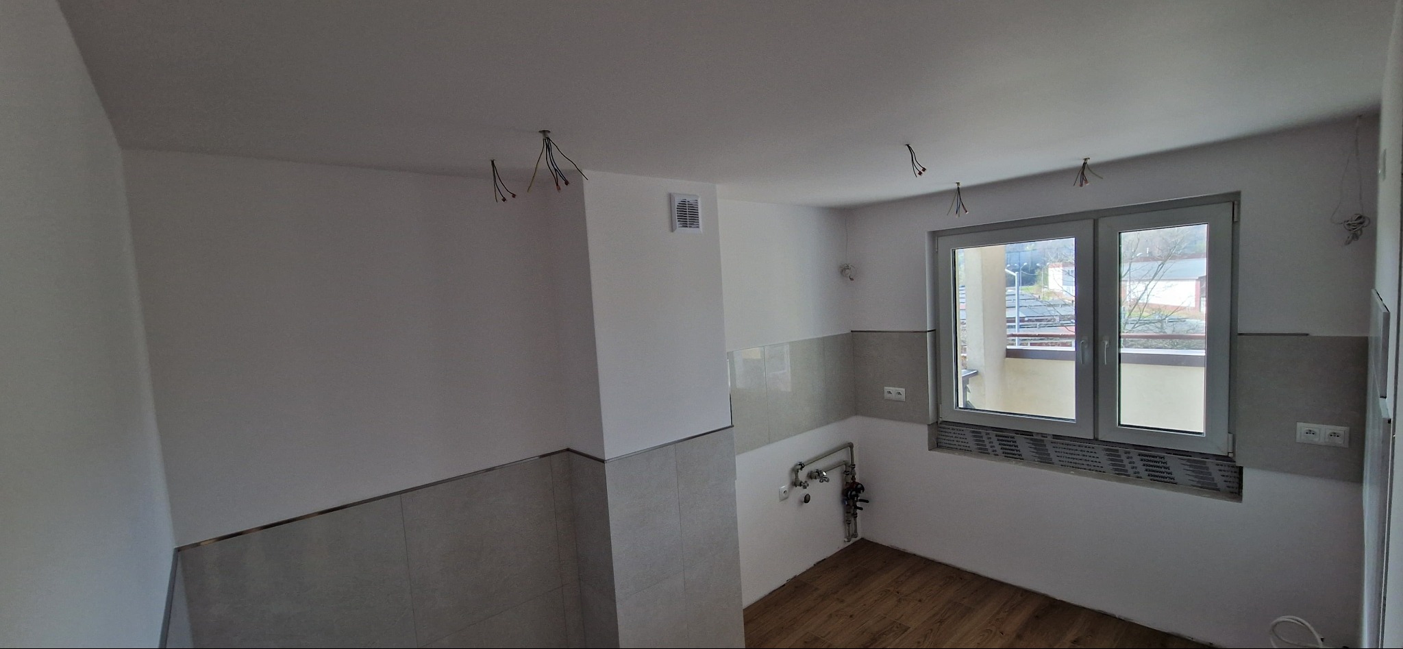 Wnętrze kuchni w trakcie częściowego wykończenia: położone płytki na ścianie, widoczne wystające kable elektryczne z sufitu, drewnopodobna podłoga, okno z widokiem na balkon.