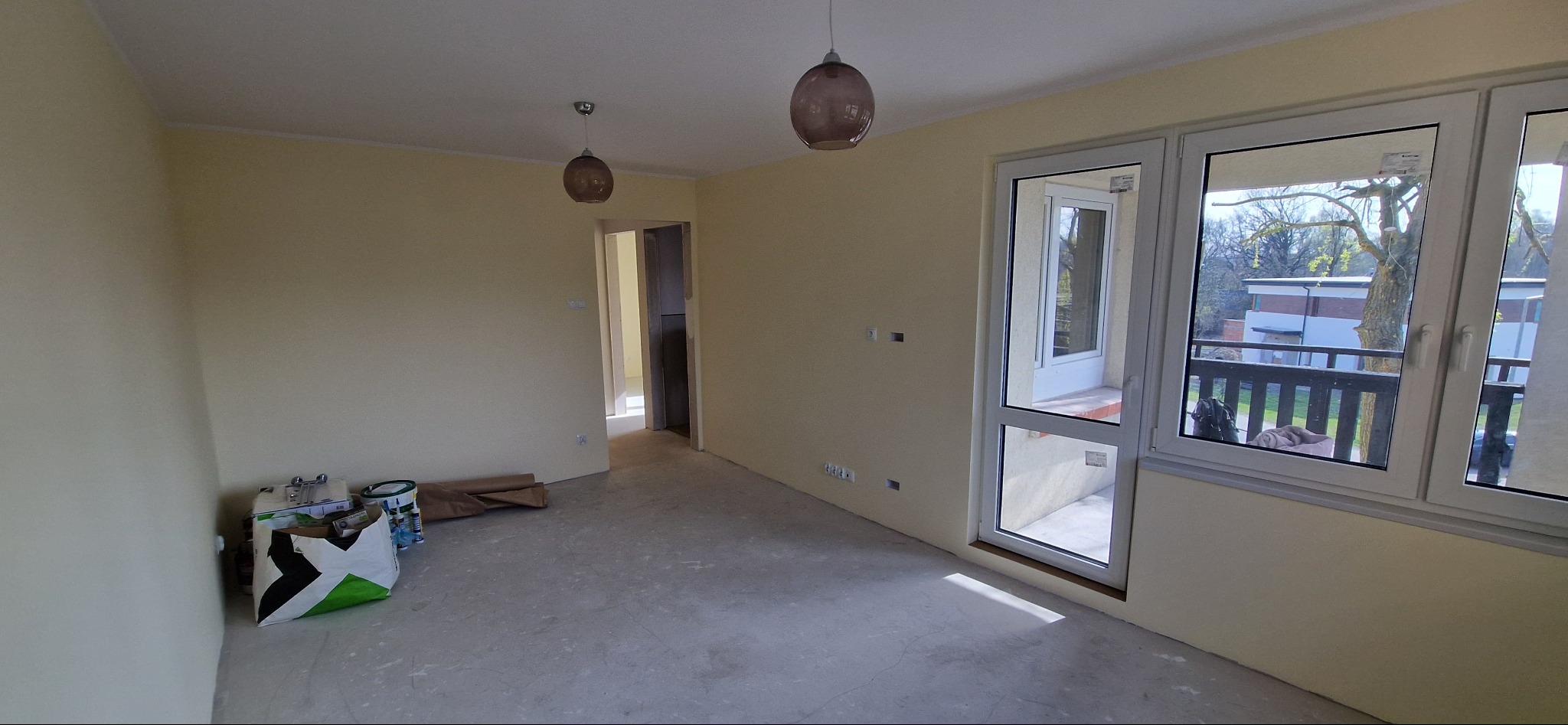 Pusty pokój w trakcie remontu z beżowymi ścianami, widoczne puszki z farbą, przez okno balkon z czarną barierką i widok na drzewo.