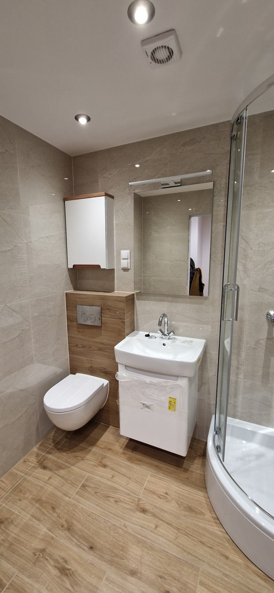 Nowoczesna łazienka w jasnych beżach z drewnopodobną podłogą, wisząca toaleta, szafka z umywalką jeszcze w folii ochronnej i kabina prysznicowa z zaokrąglonym szkłem.