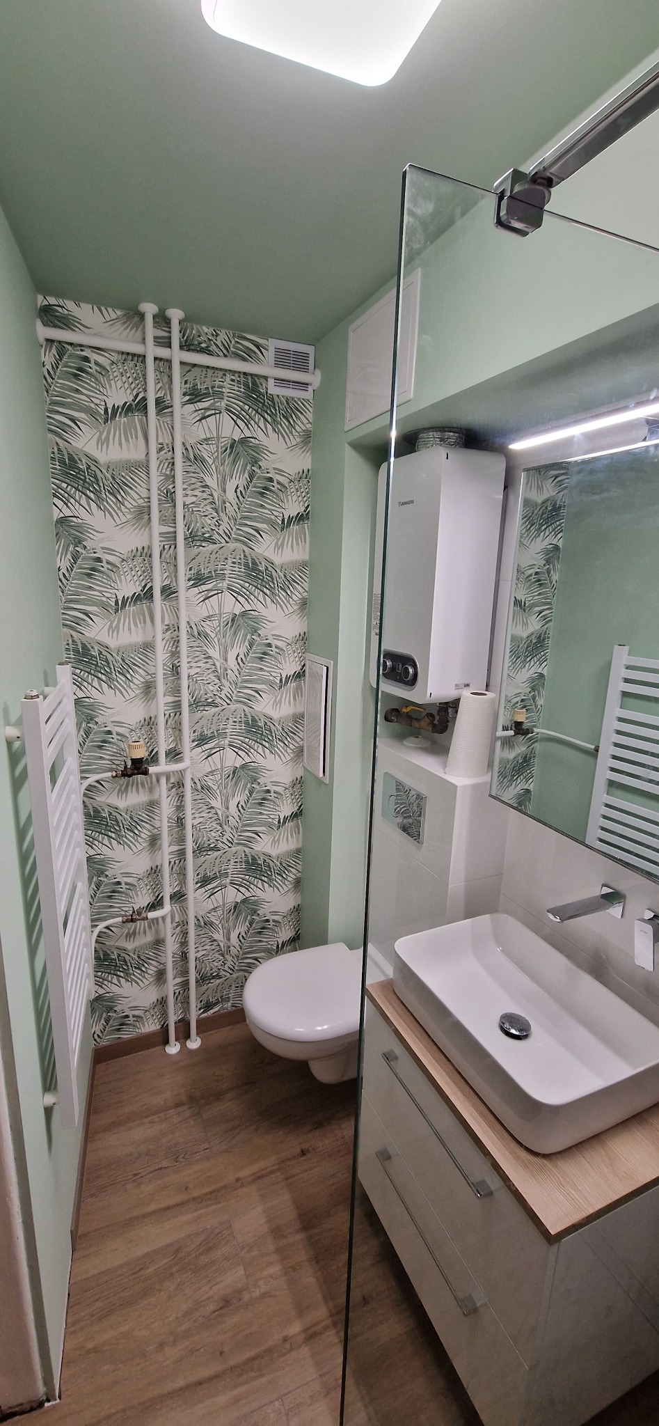 Wykończona łazienka w odcieniach zieleni z motywem tropikalnych liści na tapecie, biała umywalka nablatowa z szafką, grzejnik drabinkowy i przeszklona kabina prysznicowa.