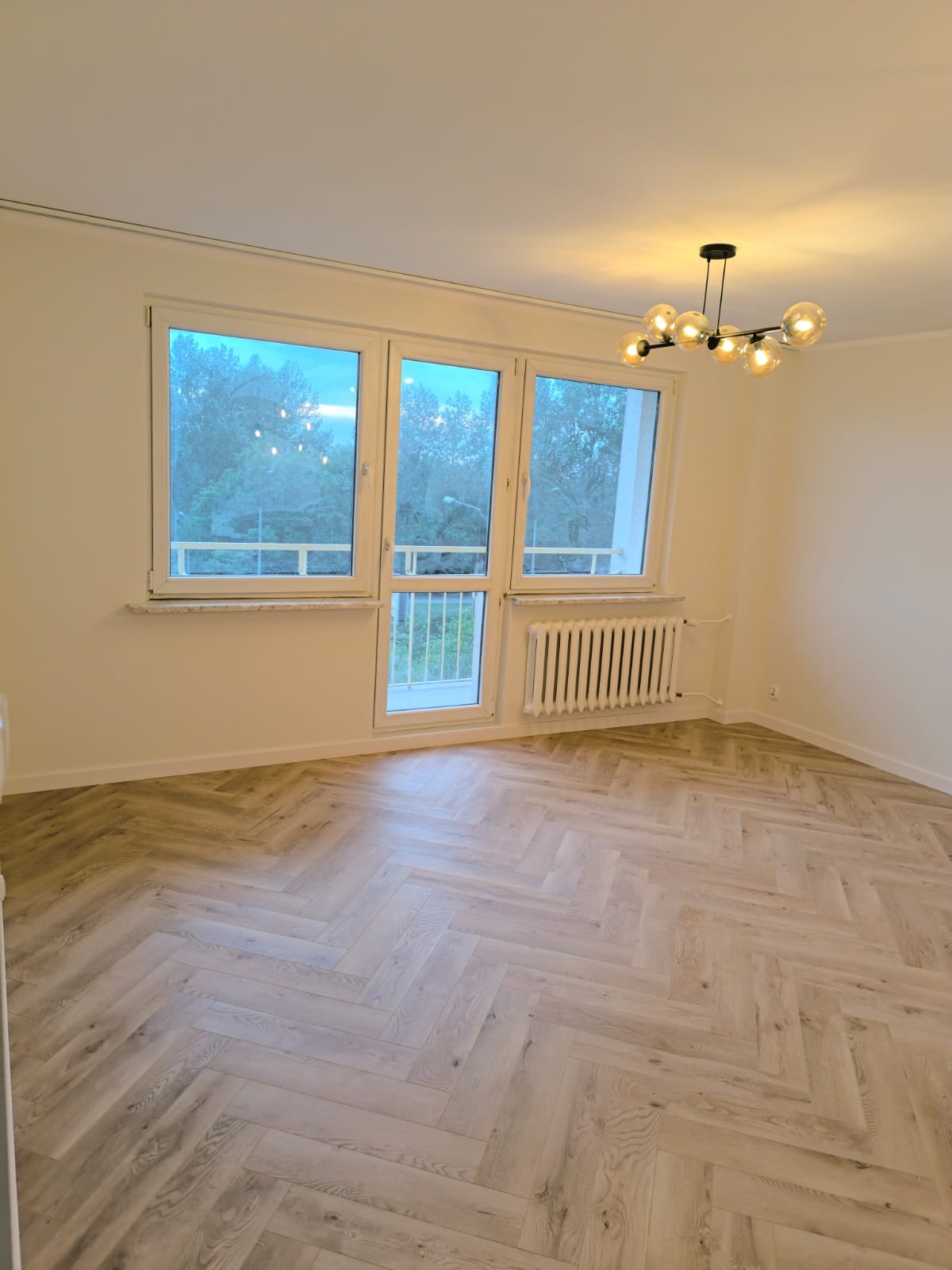 Pusty pokój po remoncie z jasną podłogą ułożoną w jodełkę, białymi ścianami, oknami z widokiem na zieleń i nowoczesną lampą sufitową z kulistymi kloszami.