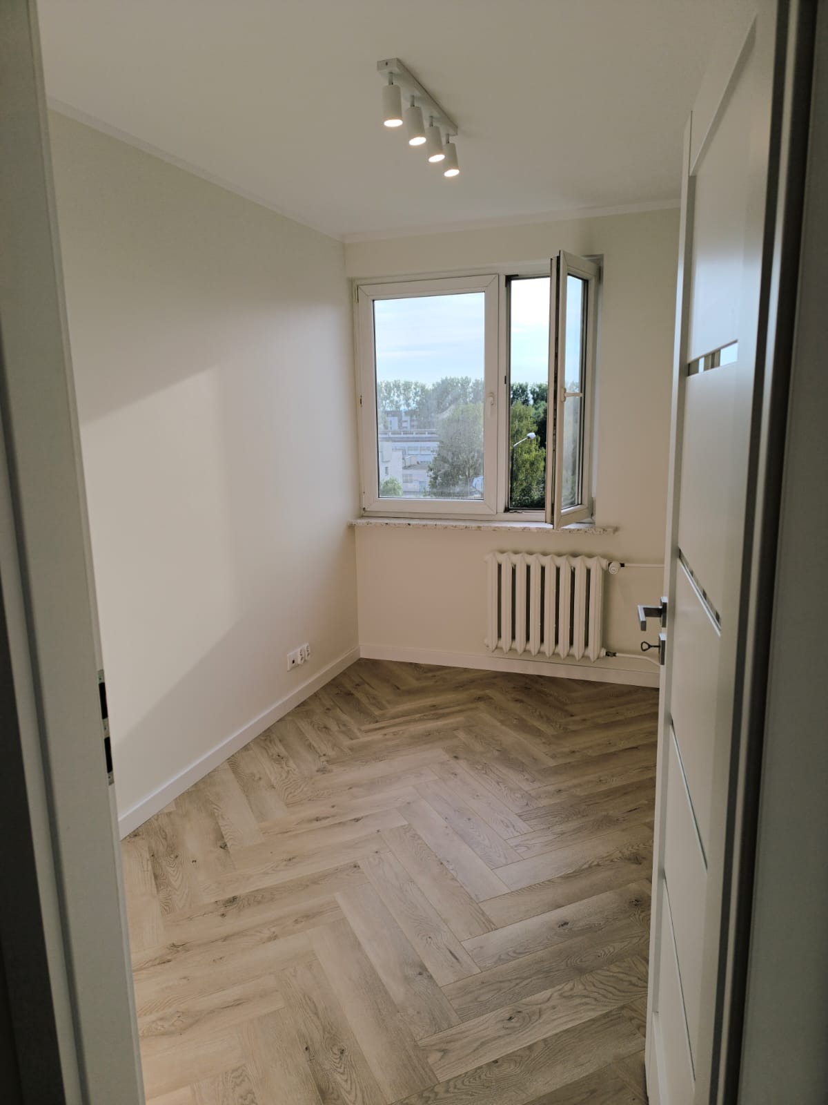 Pusty pokój z nową podłogą ułożoną w jodełkę, świeżo pomalowane ściany w odcieniach bieli, grzejnik pod oknem i nowoczesne oświetlenie sufitowe.