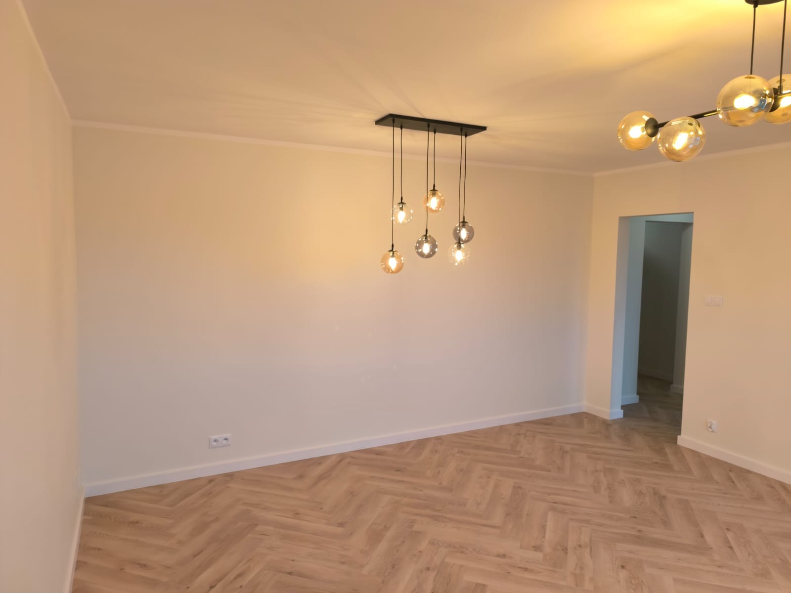 Pusty pokój z parkietem ułożonym w jodełkę, oświetlony przez dwie designerskie lampy sufitowe z kulistymi kloszami w odcieniach bursztynu i szarości.