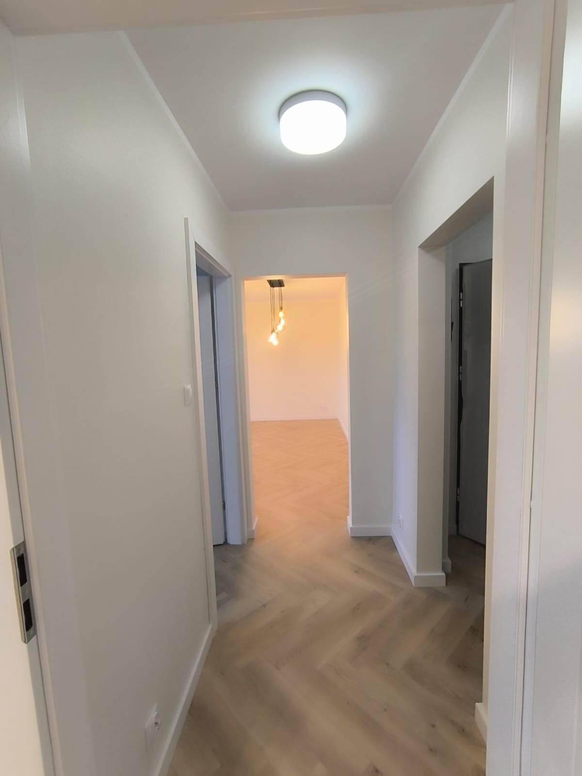 Korytarz z parkietem ułożonym we wzór jodełki, świeżo pomalowane białe ściany, widok na pokój z wiszącą lampą i białymi ścianami.
