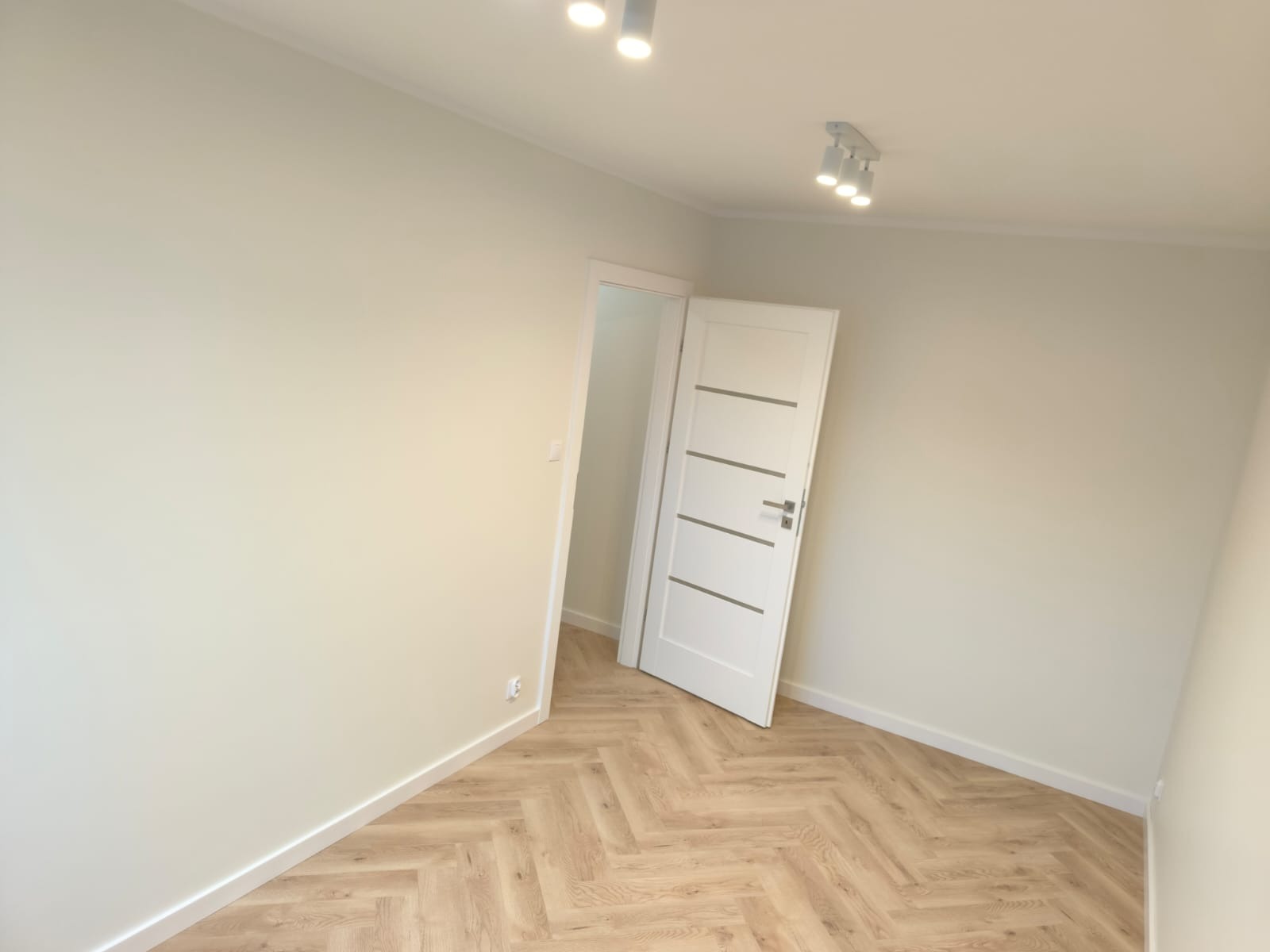 Pusty pokój z jasnymi ścianami, parkietem ułożonym w jodełkę i białą, częściowo otwartą, nowoczesną stolarką drzwiową z poziomymi, szarymi wstawkami, oświetlenie sufitowe punktowe.