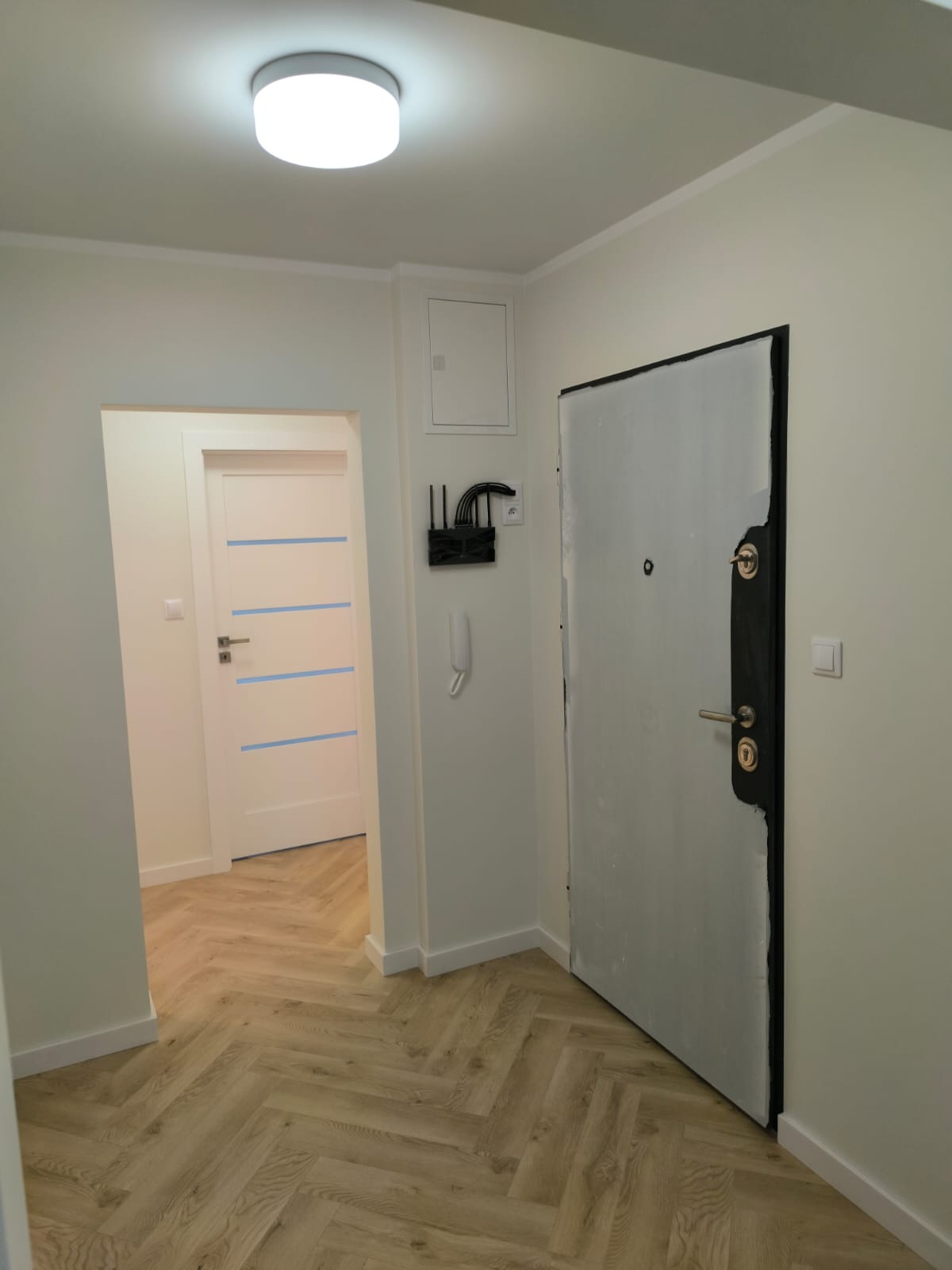 Korytarz w jasnych barwach po odświeżeniu, z parkietem ułożonym w jodełkę, białą stolarką drzwiową z niebieskimi akcentami i zabezpieczonymi folią drzwiami wejściowymi.