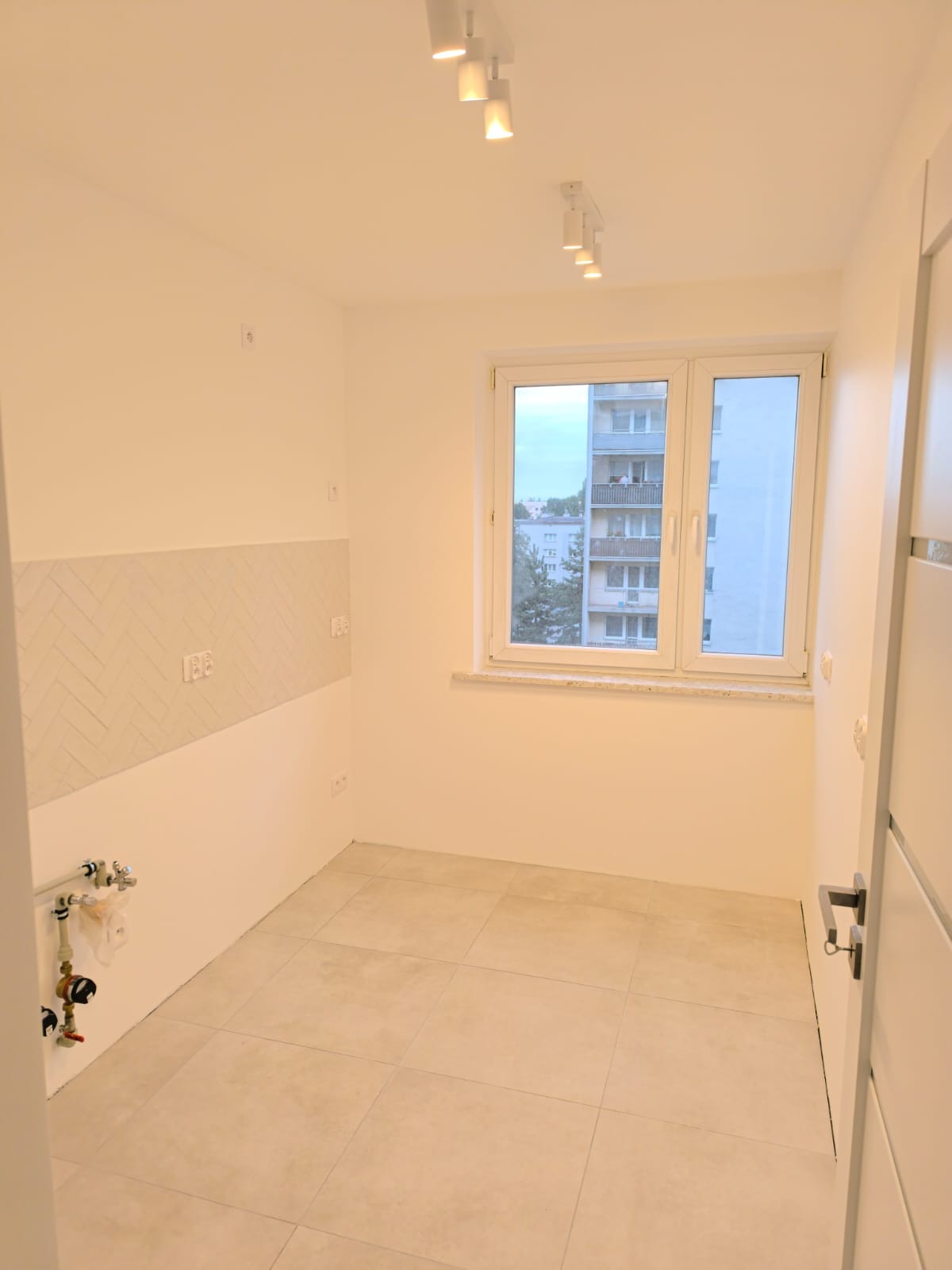 Jasne, puste pomieszczenie po remoncie z beżowymi płytkami podłogowymi i fragmentem ściany wyłożonej płytkami w jodełkę, widoczne rury instalacji wodnej i okno z widokiem na blok.