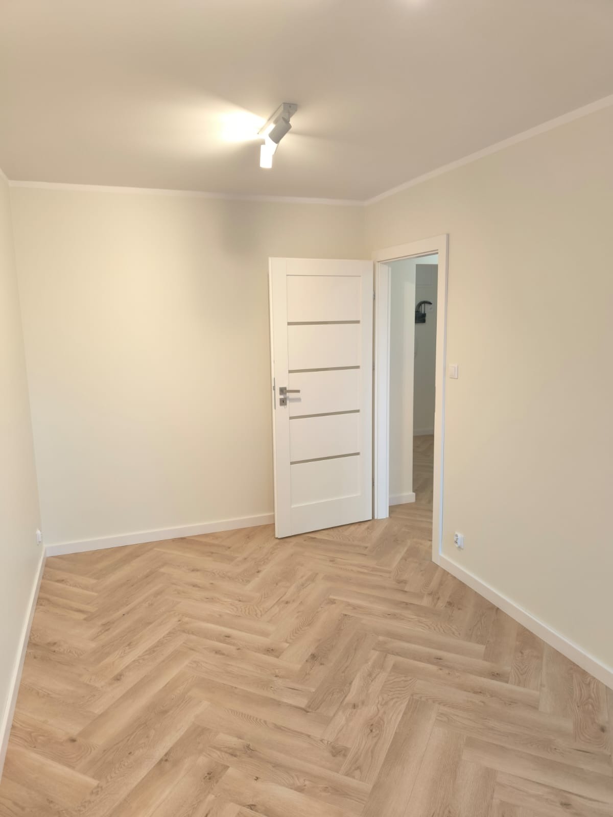Pusty pokój z parkietem ułożonym w jodełkę, świeżo pomalowanymi ścianami w odcieniach beżu i białymi drzwiami wewnętrznymi z poziomymi frezowaniami.