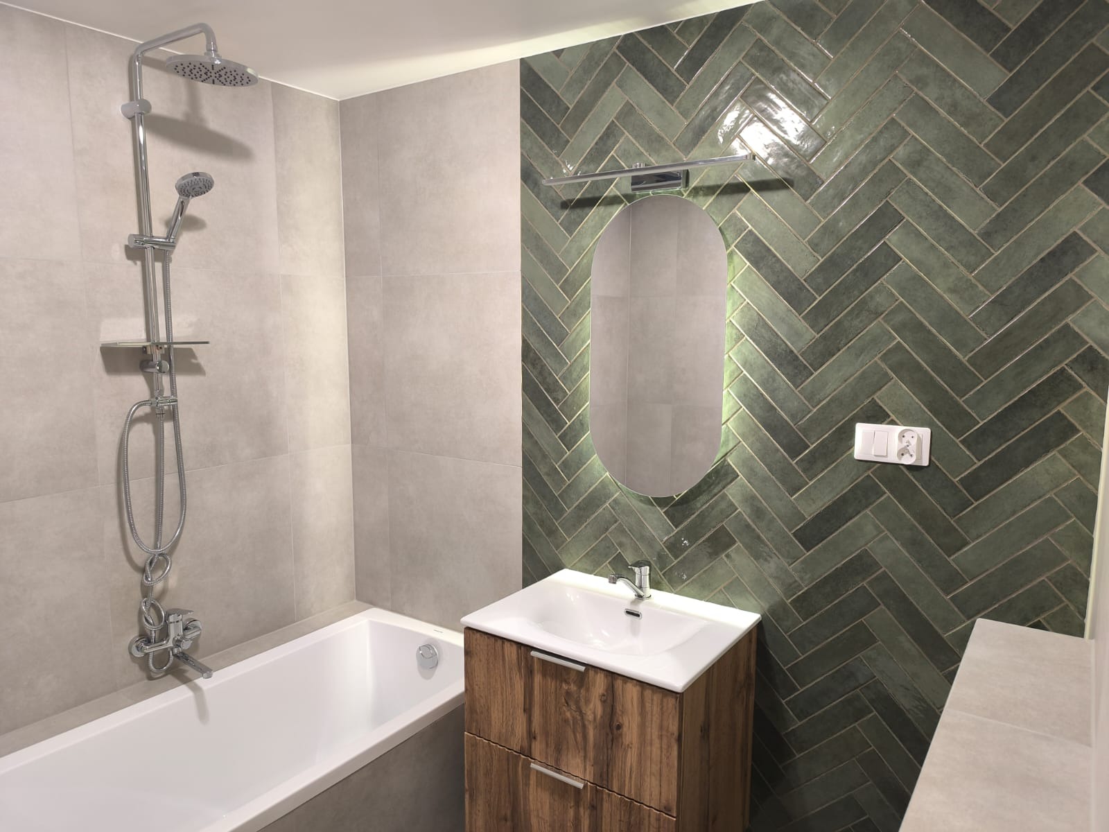 Nowoczesna łazienka z zielonymi płytkami ułożonymi w jodełkę, podświetlanym owalnym lustrem nad drewnianą szafką umywalkową i białą wanną z chromowaną armaturą.