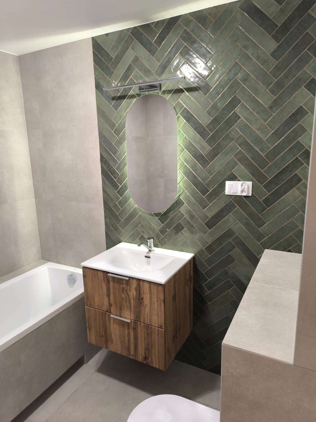 Nowoczesna łazienka z zielonymi płytkami ułożonymi w jodełkę, drewniana szafka pod umywalką z owalnym lustrem podświetlanym LED i białą wanną.