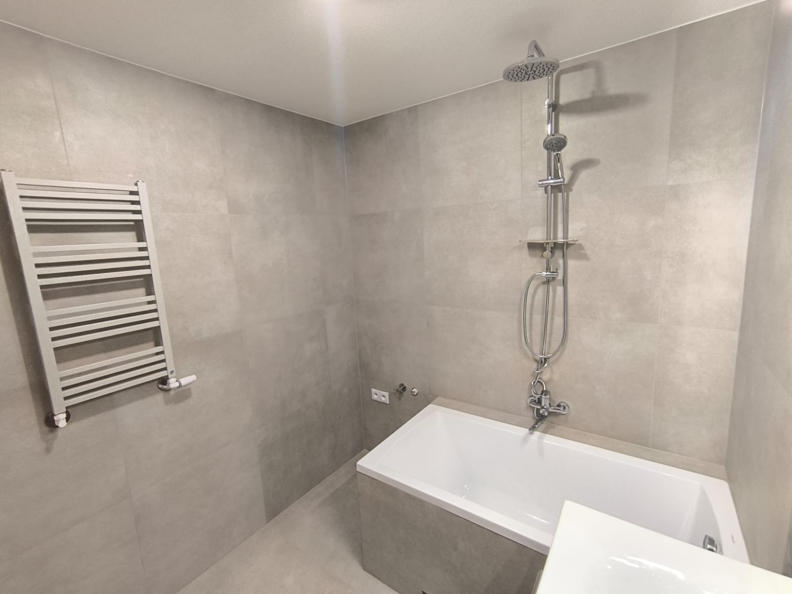 Wykończona łazienka w szarościach, z białą wanną i grzejnikiem drabinkowym; widoczny zestaw prysznicowy z deszczownicą i bateria wannowa.