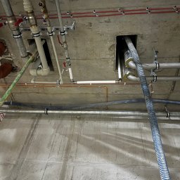 SDV GROUP - Widok instalacji pod stropem w Krakowie: rury różnego typu (metalowe, plastikowe, karbowane) i średnicy, zawory, mocowania, izolacja termiczna na rurach, surowy beton.