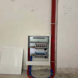 SDV GROUP - Rozdzielacz ogrzewania podłogowego w białej szafce, z czerwonymi i niebieskimi rurami, przy ścianie z notatkami i prowizoryczną żarówką.