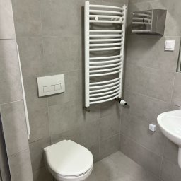 SDV GROUP - Nowoczesna łazienka z szarymi płytkami, wiszącą toaletą, białą drabinką grzejnikową z termostatem, podajnikiem na ręczniki papierowe i umywalką.