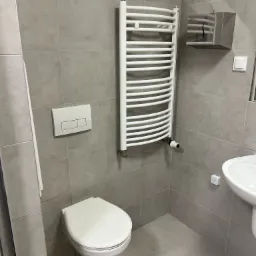 Nowoczesna łazienka z szarymi płytkami, wiszącą toaletą, białą drabinką grzejnikową z termostatem, podajnikiem na ręczniki papierowe i umywalką.