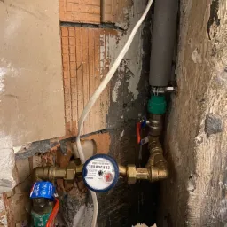 Zamknięty wnęką wodomierz z zaworami odcinającymi i rurami miedzianymi, widoczne uszkodzenia tynku i cegły na ścianie.