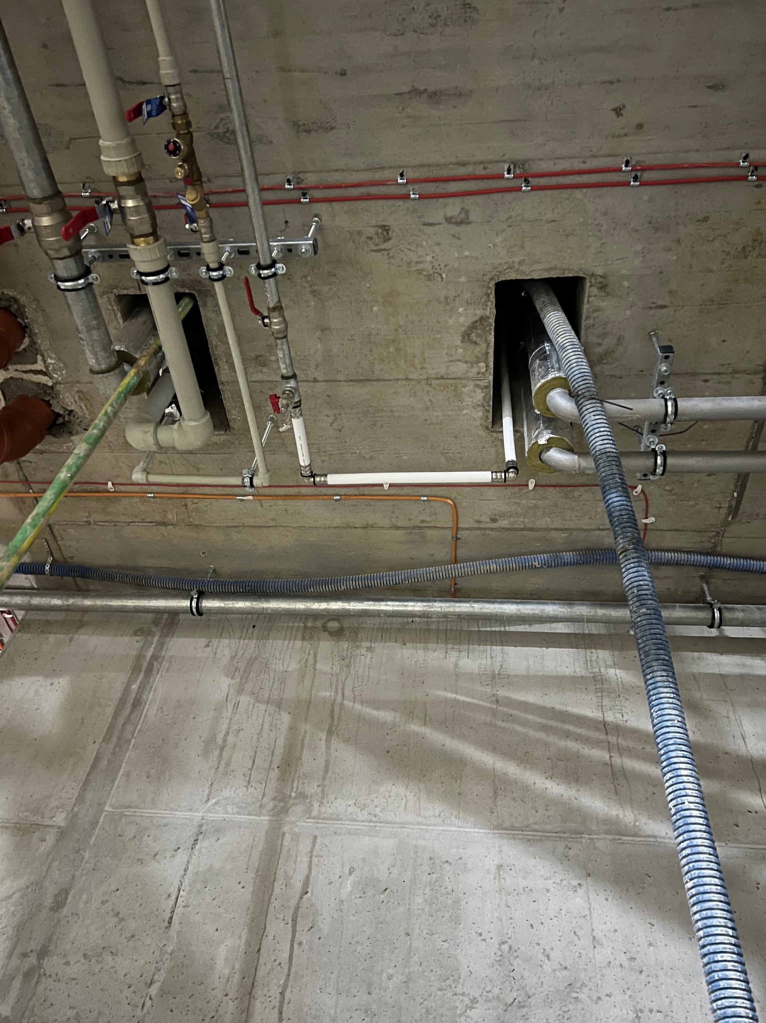 Widok instalacji pod stropem w Krakowie: rury różnego typu (metalowe, plastikowe, karbowane) i średnicy, zawory, mocowania, izolacja termiczna na rurach, surowy beton.