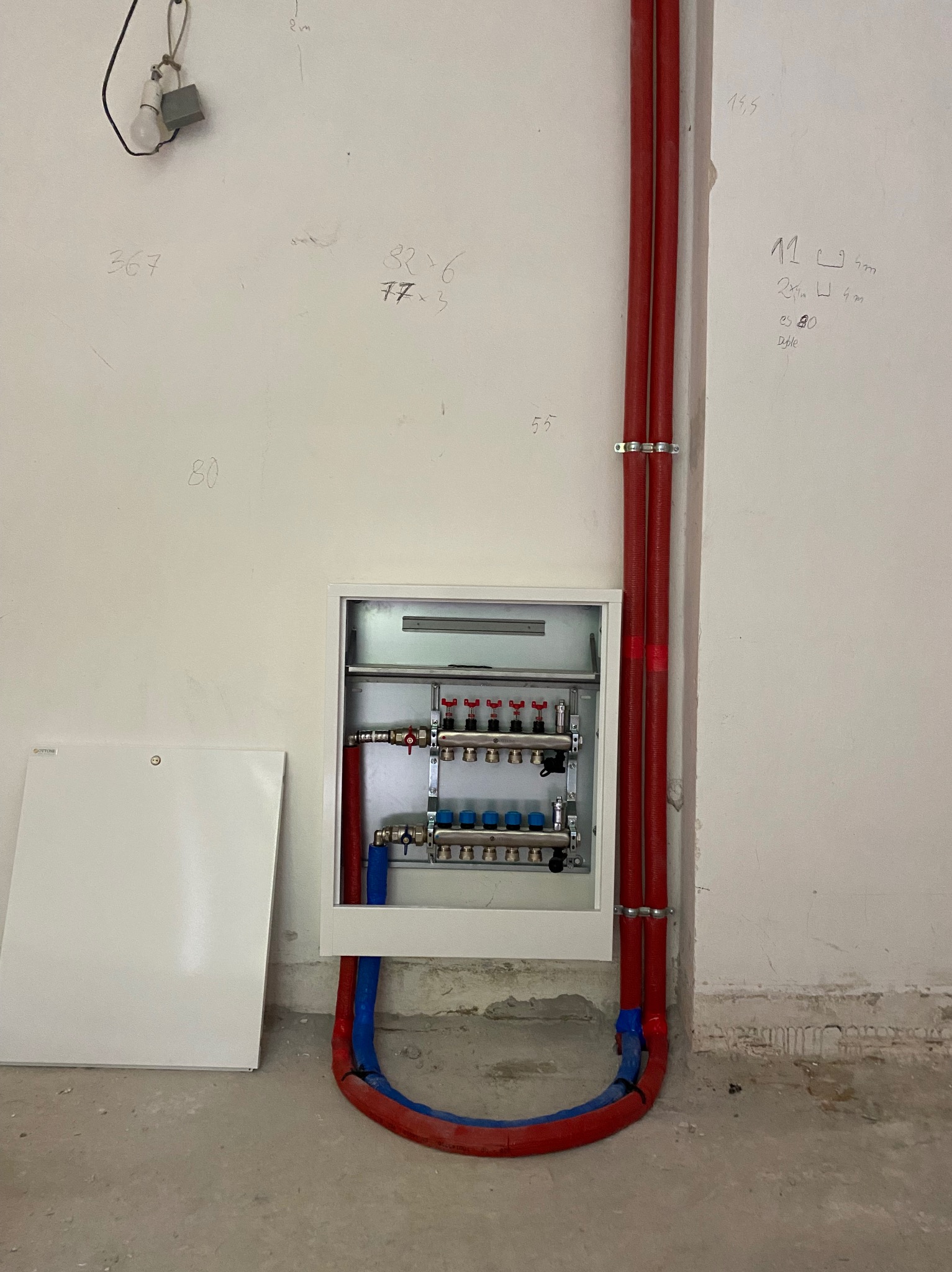 Rozdzielacz ogrzewania podłogowego w białej szafce, z czerwonymi i niebieskimi rurami, przy ścianie z notatkami i prowizoryczną żarówką.