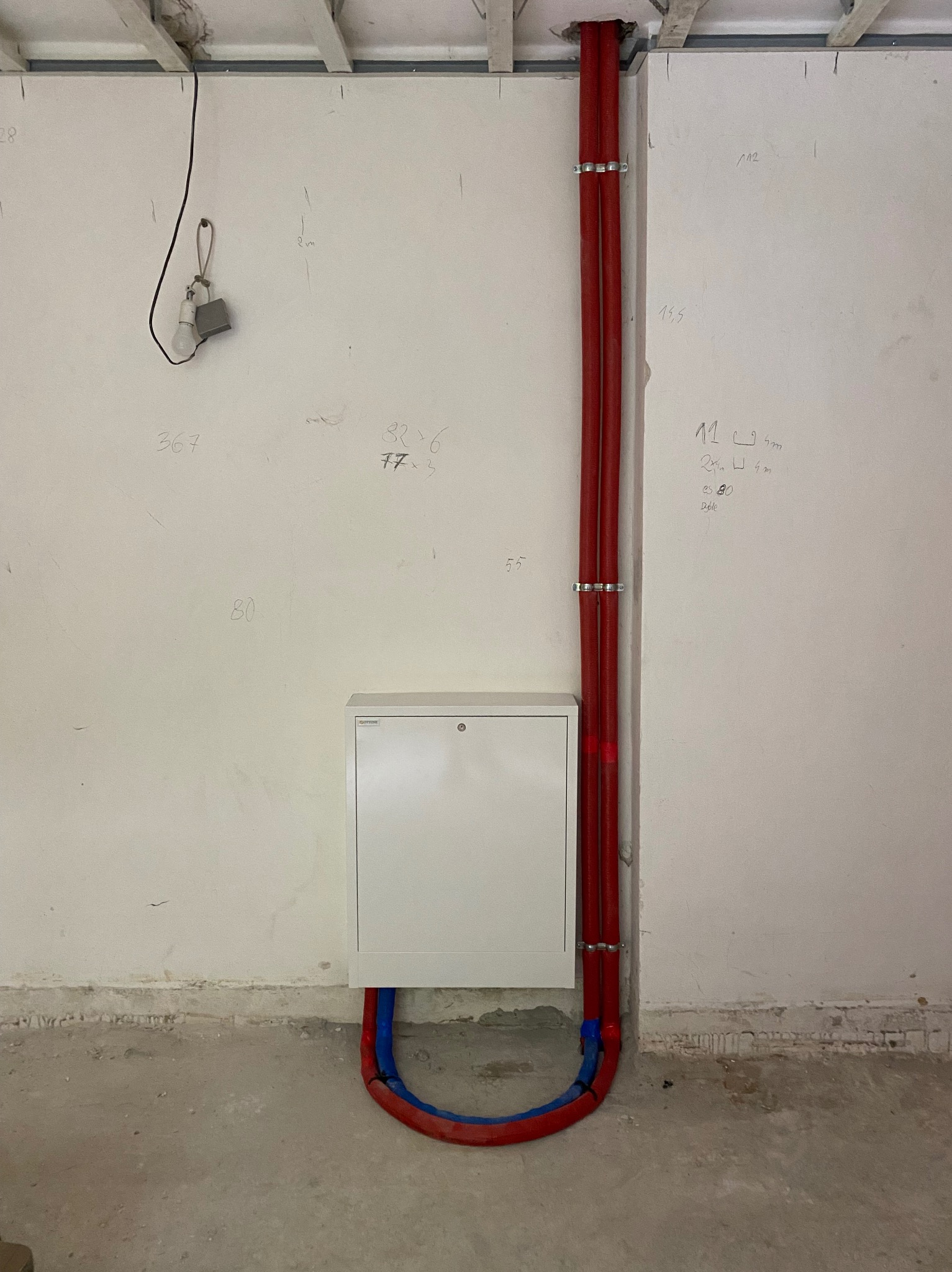 Biała skrzynka instalacyjna na ścianie w surowym wnętrzu, z podłączonymi czerwonymi rurami, zabezpieczonymi srebrnymi klamrami, oraz fragmentem niebieskiej rury; na ścianie widoczne odręczne notatki.