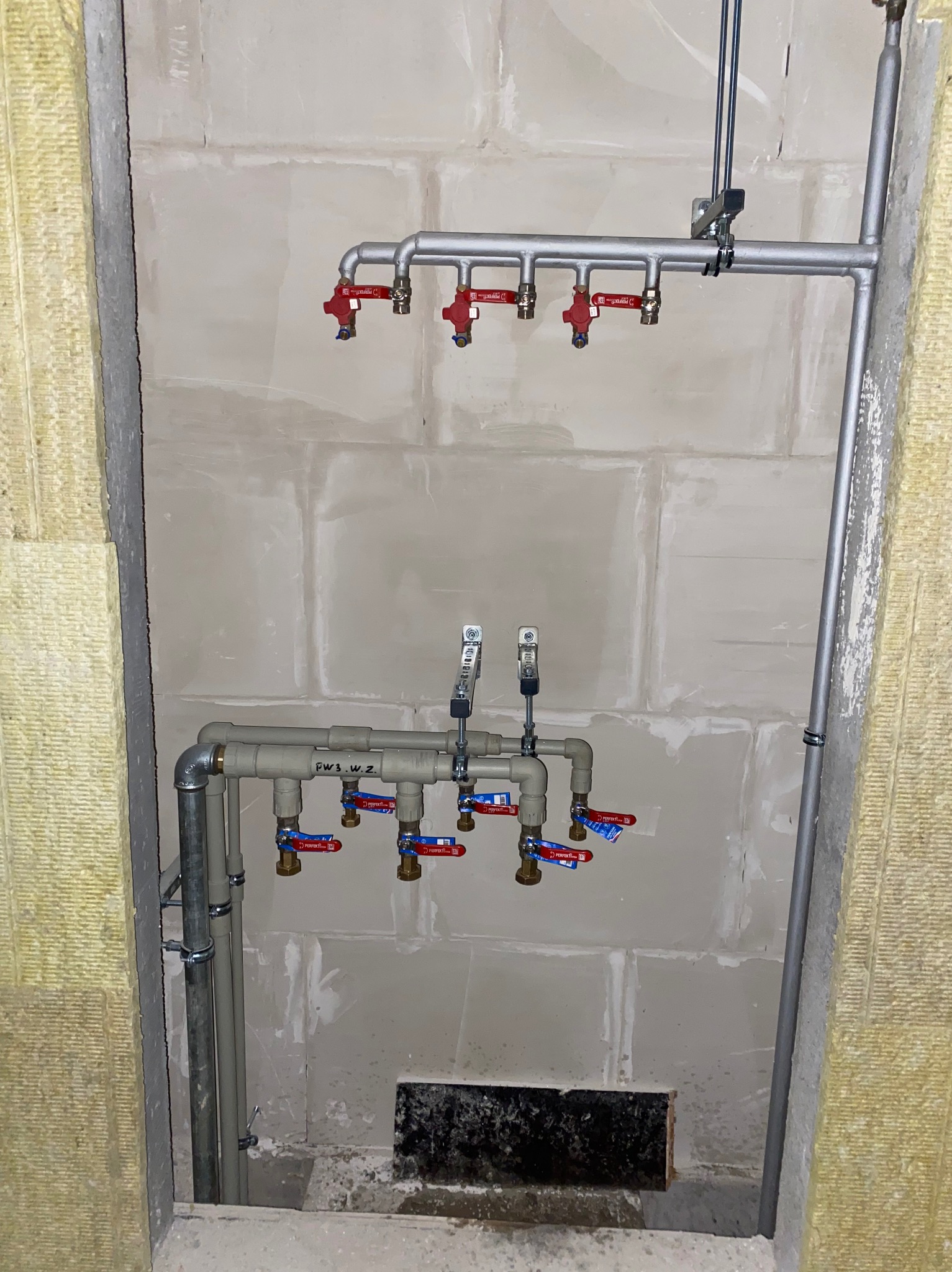 Rozdzielacz instalacji wodnej z czerwonymi zaworami, zamontowany na surowej ścianie z izolacją z wełny mineralnej, widok na fragment instalacji w trakcie montażu.