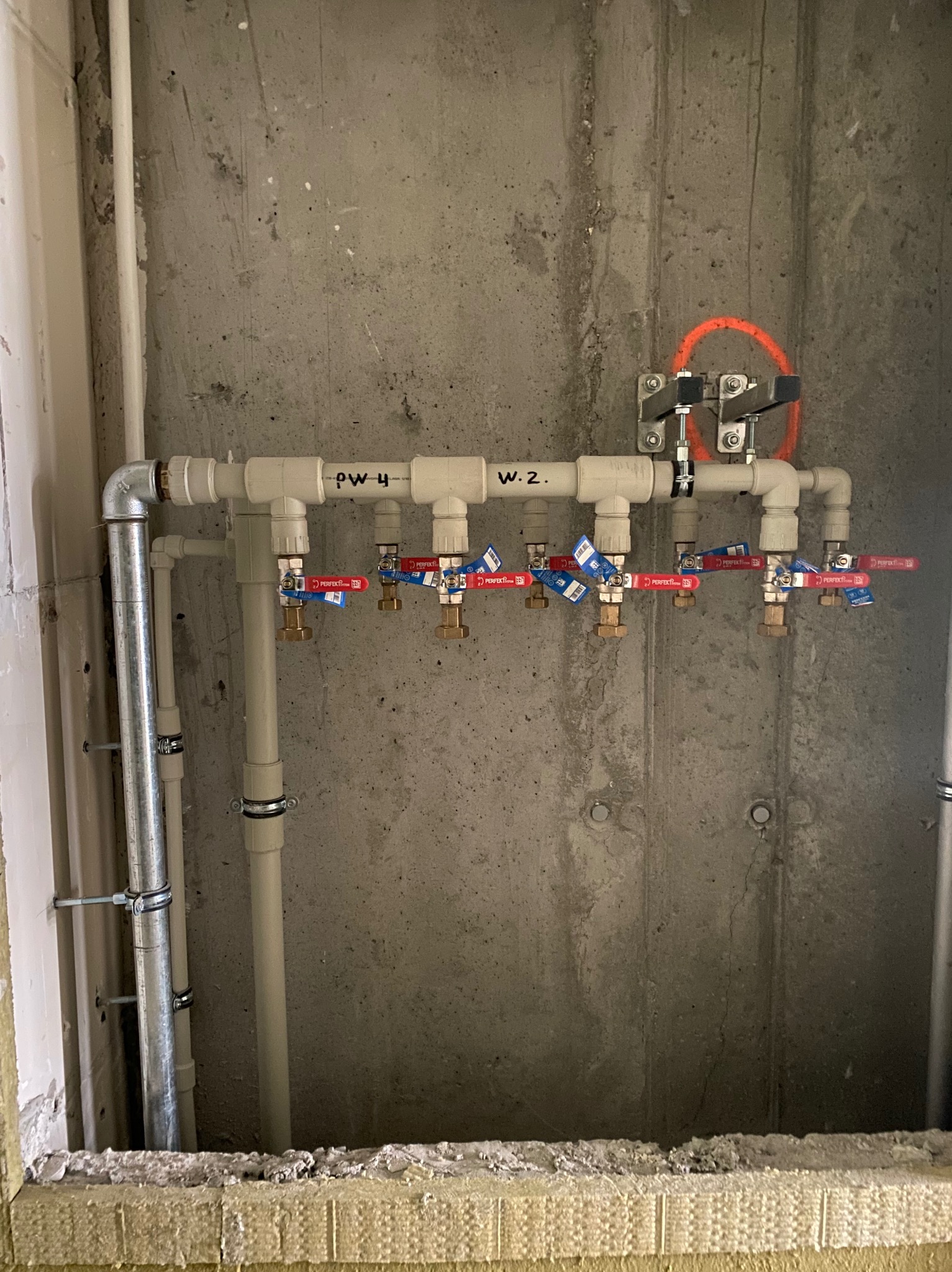 Rozdzielacz instalacji wodnej z czterema zaworami odcinającymi Perfect z czerwonymi rączkami i niebieskimi etykietami, zamontowany na surowej betonowej ścianie z pomarańczowym okręgiem, rury...