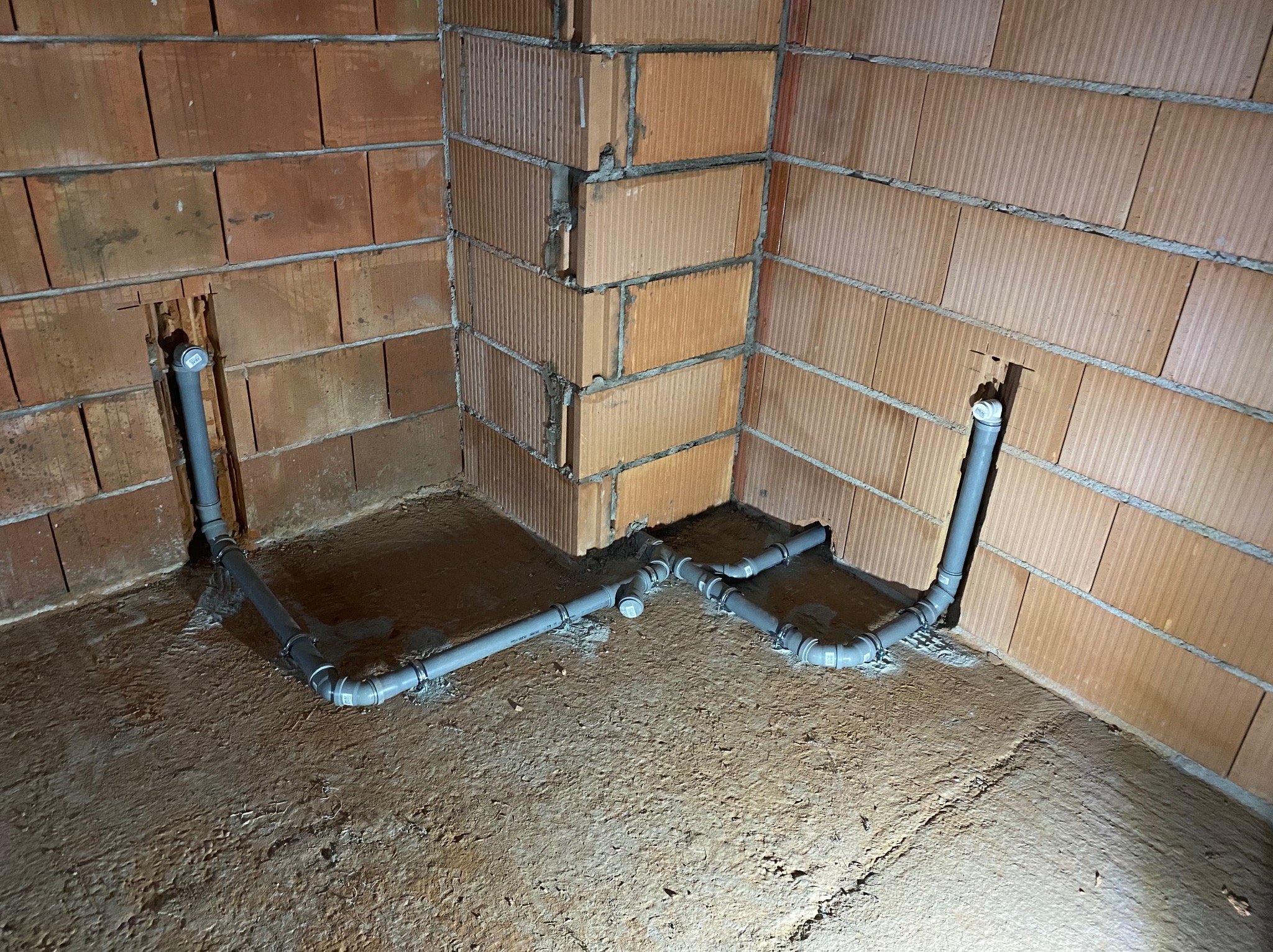 Szare rury instalacji wodnej w surowym stanie, mocowane do ściany z czerwonej cegły dziurawki, widoczne obejmy mocujące, betonowa podłoga w trakcie wylewki.