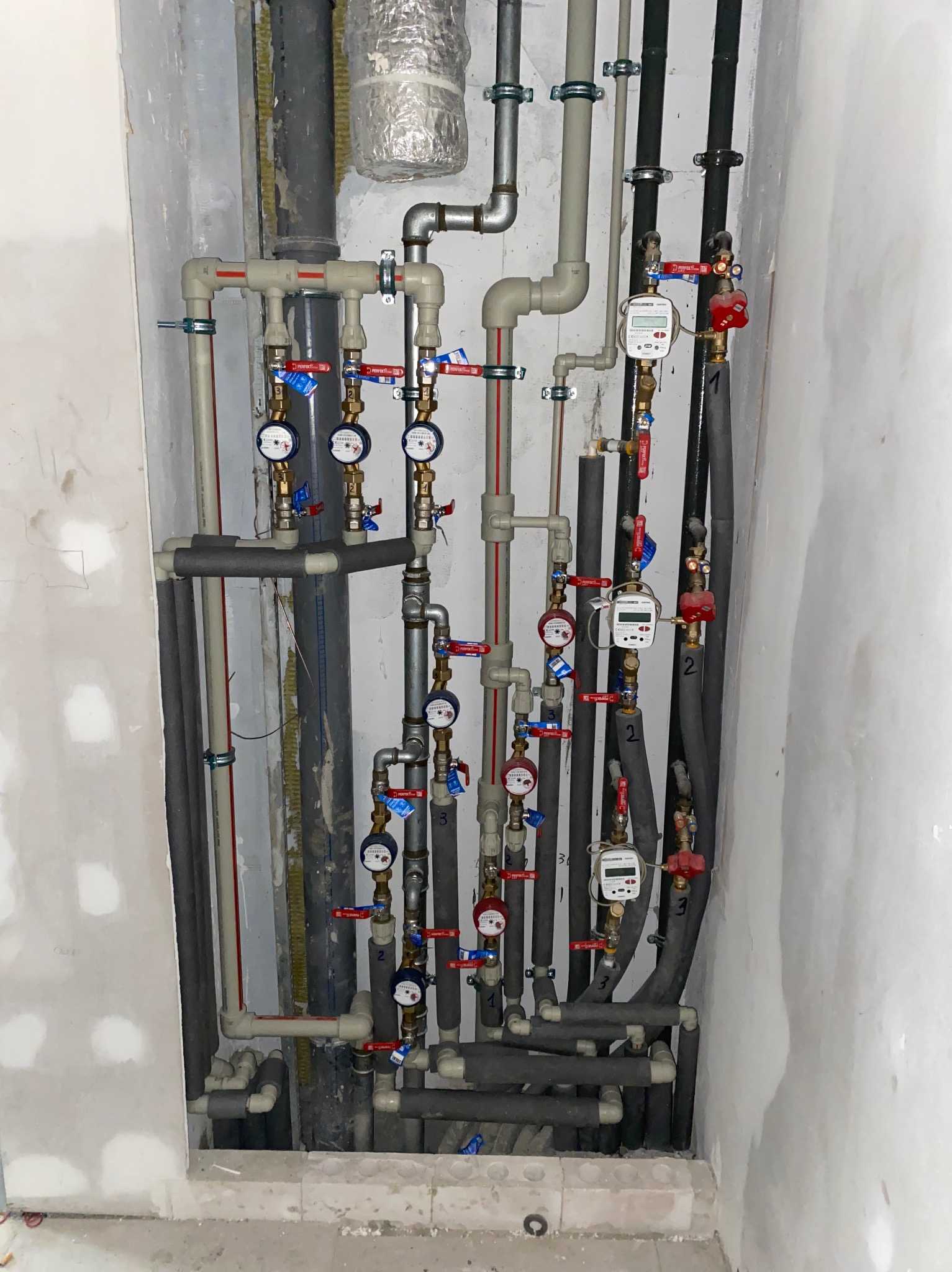Zestawienie rur instalacyjnych z zaworami i licznikami, widoczne różne materiały i izolacje, montaż w surowym stanie, ściany w trakcie wykańczania.