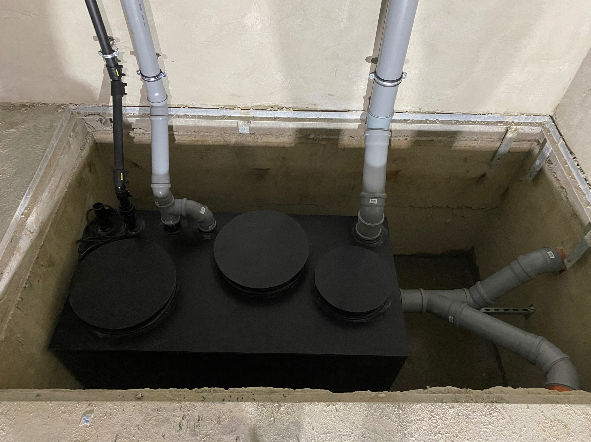 Czarna skrzynka z trzema okrągłymi pokrywami w betonowej studzience, połączona z szarymi rurami PCV z licznymi złączkami i obejmami mocującymi do ściany i sufitu.