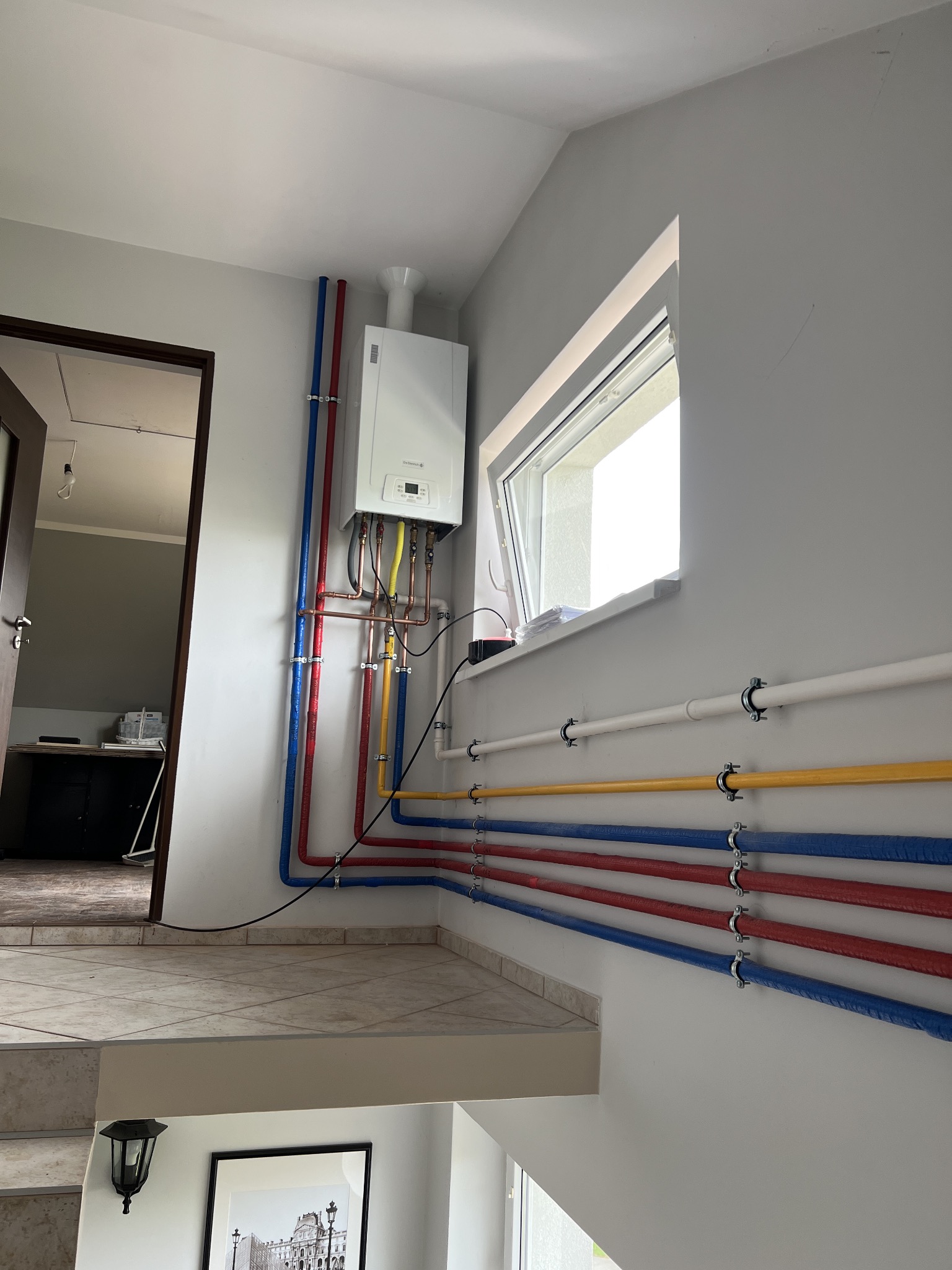 Nowoczesny, wiszący piec gazowy De Dietrich z kolorowymi rurami instalacji grzewczej (czerwone, niebieskie, żółte) zamontowany na jasnej ścianie przy oknie, widok z dołu schodów.