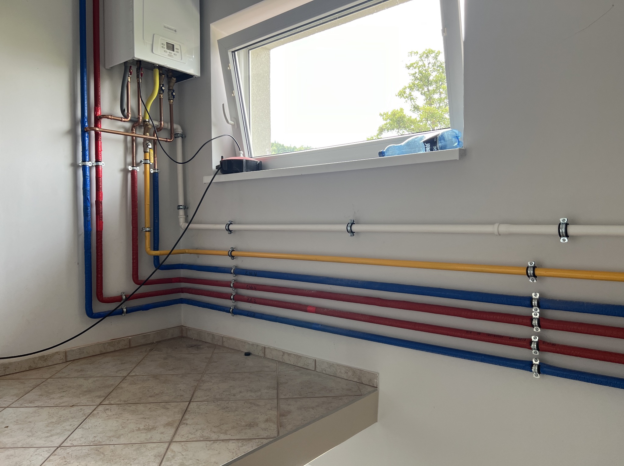 Nowa instalacja grzewcza z kotłem gazowym De Dietrich i kolorowymi rurami (czerwone, niebieskie, żółte) zamocowanymi do ściany za pomocą metalowych uchwytów, widok na okno z butelką wody.