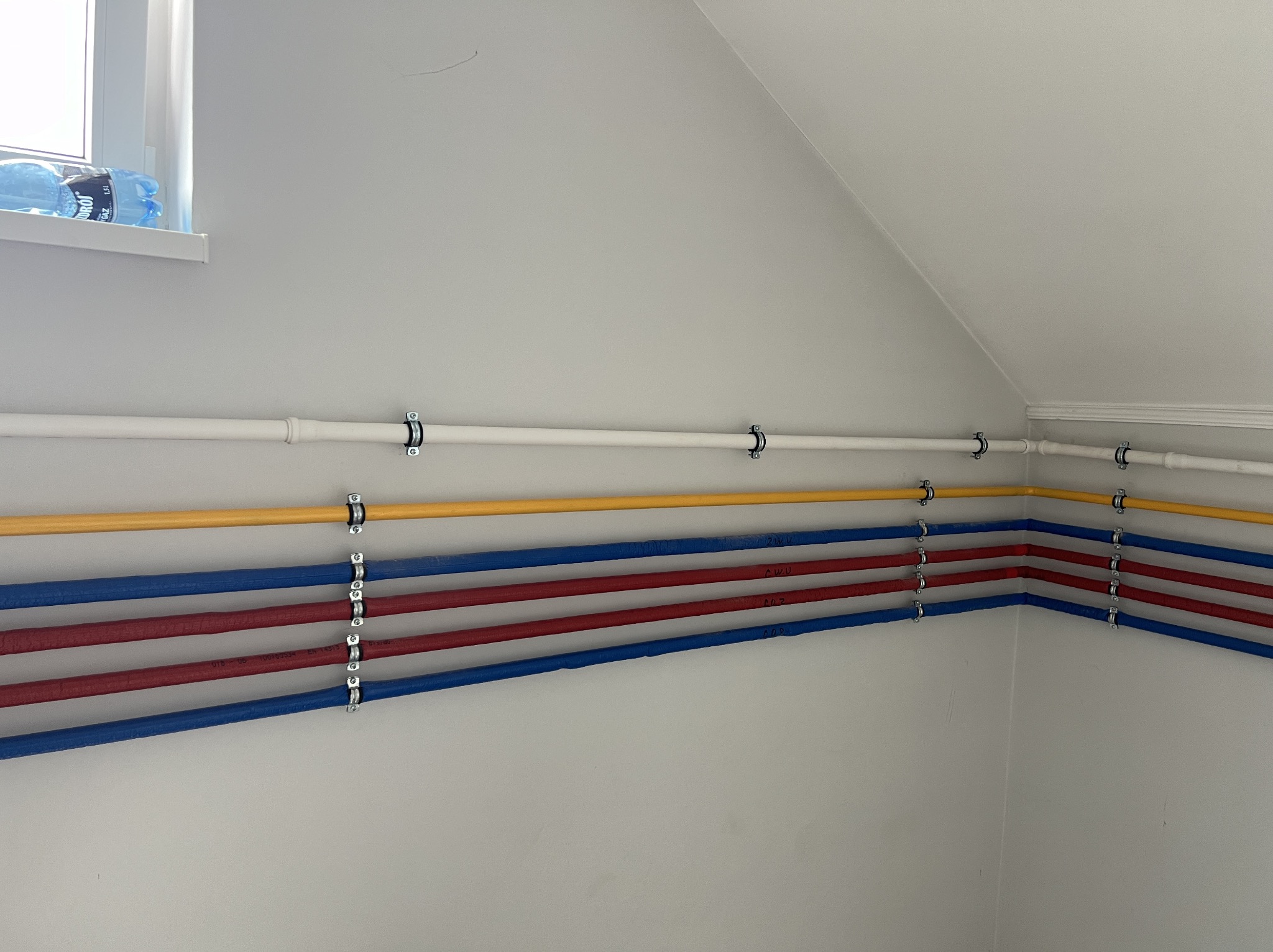 Rzędy kolorowych rur (biała, żółta, czerwona, niebieska) przymocowane do ściany za pomocą metalowych uchwytów. Widoczne napisy na rurach. Butelka wody na parapecie okna w tle.