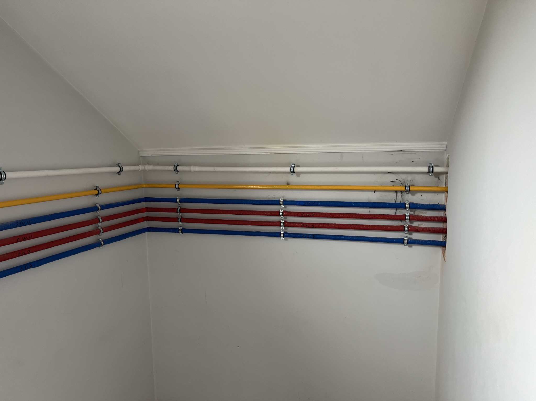 Wewnętrzne ułożenie rur instalacyjnych w kolorach białym, żółtym, czerwonym i niebieskim, przymocowanych metalowymi uchwytami do białej ściany w pomieszczeniu ze skośnym sufitem.