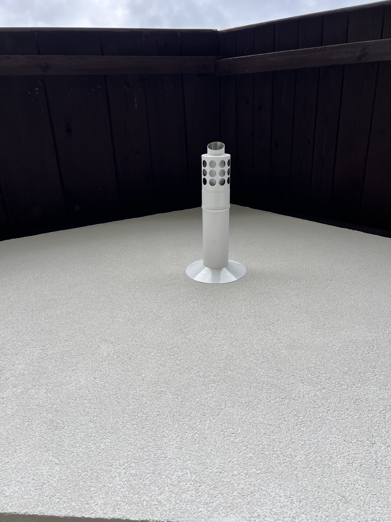 Biały kominek wentylacyjny z perforacją, zamontowany na jasnym, teksturowanym dachu, z widoczną brązową deską okapową w tle.