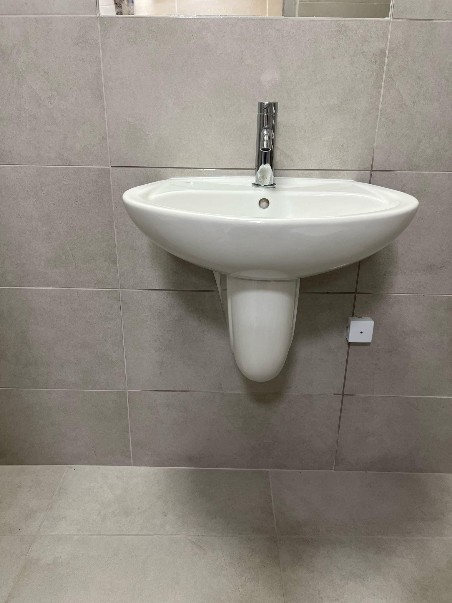 Biała umywalka zamontowana na ścianie z szarych płytek, z chromowaną baterią i białym syfonem. Widoczny biały włącznik światła po prawej stronie umywalki.